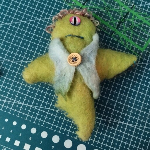 A green creature with one eye, made of felt, with curly wool hair and a smart looking wool vest.